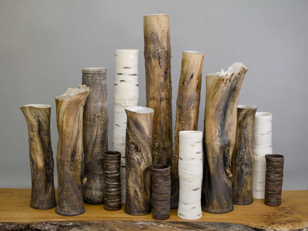 A group of ceramic tree trunk vessels by Sonya Wilkins Ceramics gathered together as a group like a copse of trees against a grey background, sitting on an oak wood shelf
