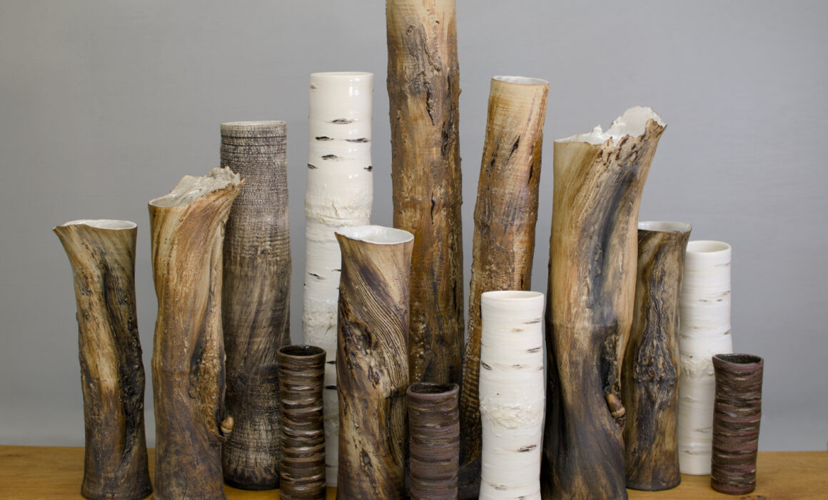 A group of ceramic tree trunk vessels by Sonya Wilkins Ceramics gathered together as a group like a copse of trees against a grey background, sitting on an oak wood shelf