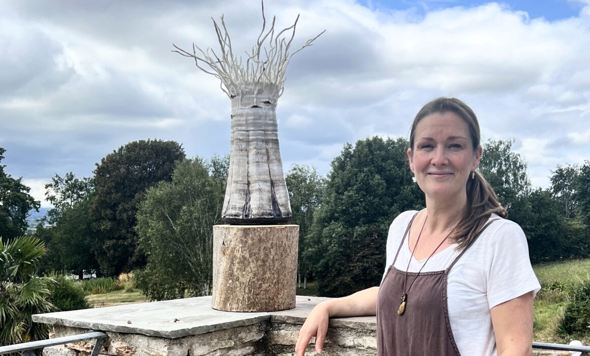 Sonya Wilkins stood next to her ceramic silver birch tree vessel exhibit on the patio of Stoberry Park Gardens with a cloud and blue sky backdrop and distant trees