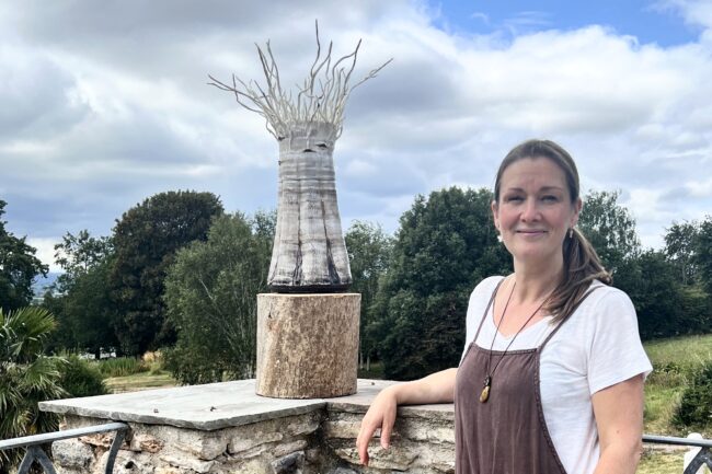 Sonya Wilkins stood next to her ceramic silver birch tree vessel exhibit on the patio of Stoberry Park Gardens with a cloud and blue sky backdrop and distant trees