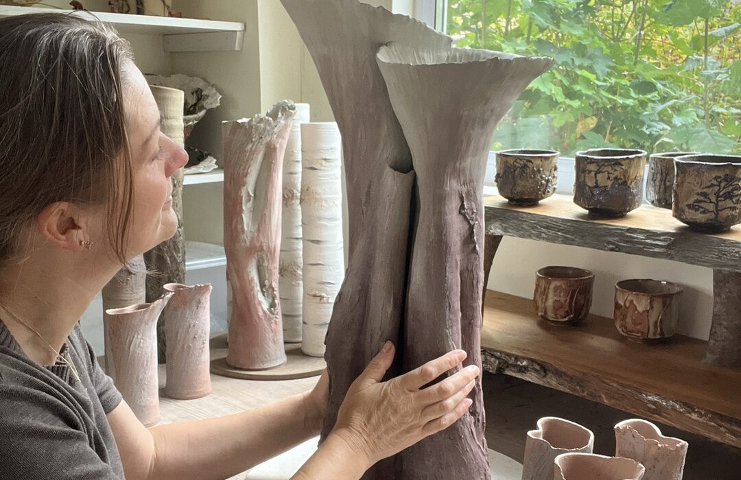 Sonya Wilkins touching a clay tree vessel in her studio, connecting with the visceral material and intention of making; allowing her passion for clay to be transmuted. In the background, there are many other clay vessels surrounding her, all creations from her love of nature and clay