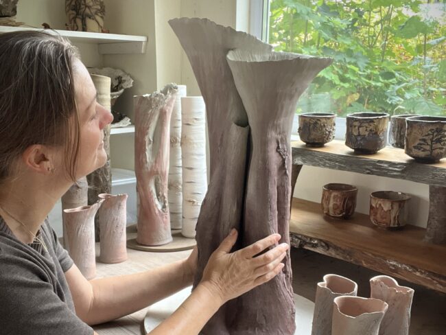 Sonya Wilkins touching a clay tree vessel in her studio, connecting with the visceral material and intention of making; allowing her passion for clay to be transmuted. In the background, there are many other clay vessels surrounding her, all creations from her love of nature and clay