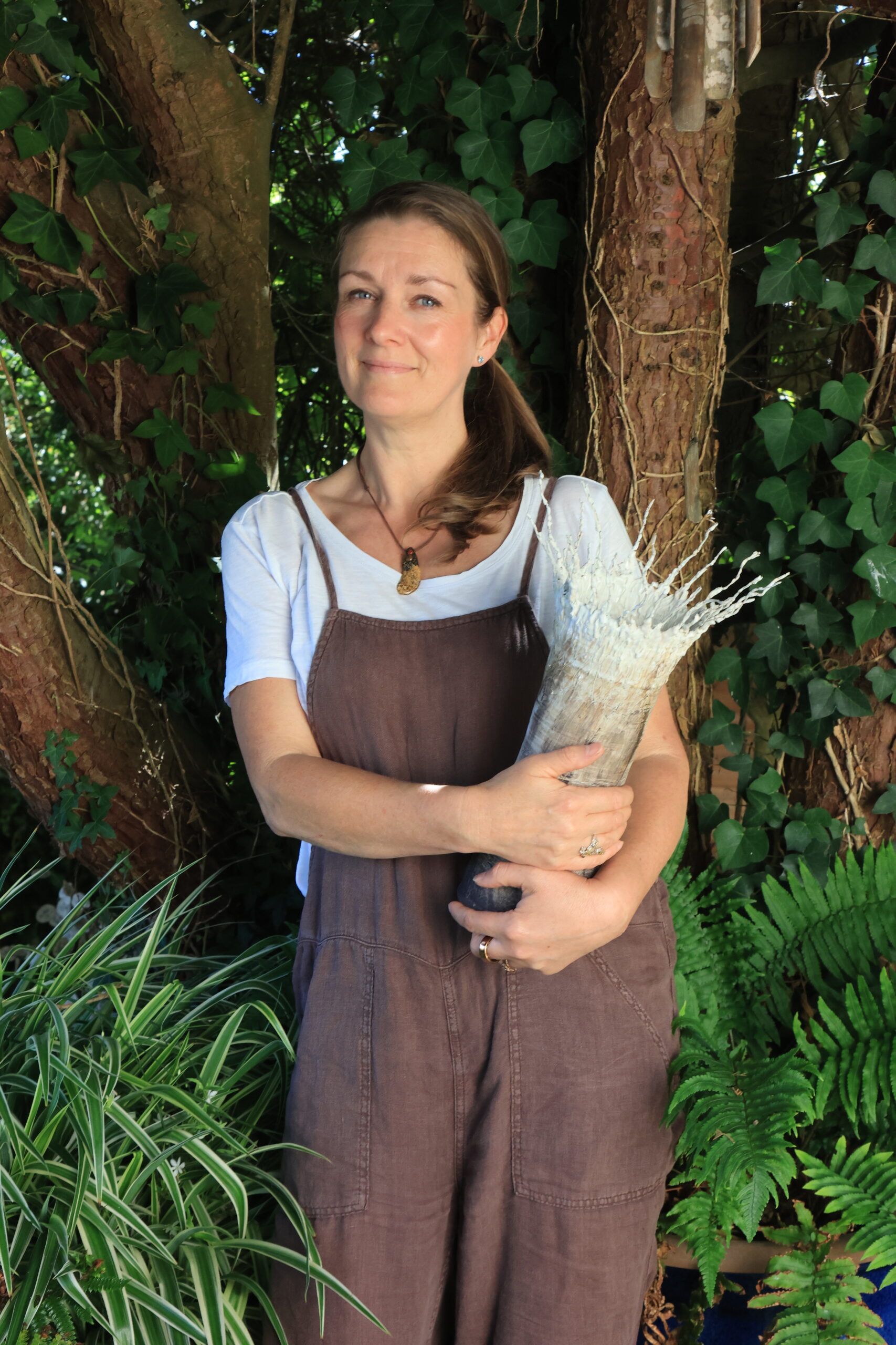 Sonya Wilkins holding one of her ceramic tree vessel creations, against a backdrop pine tree trunk, ivy and ferns