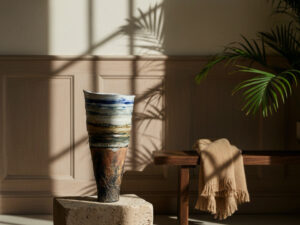 Ceramic layered landscape vessel sat on a stone plinth within an interior design environment with a plant, low table and blanket in the background